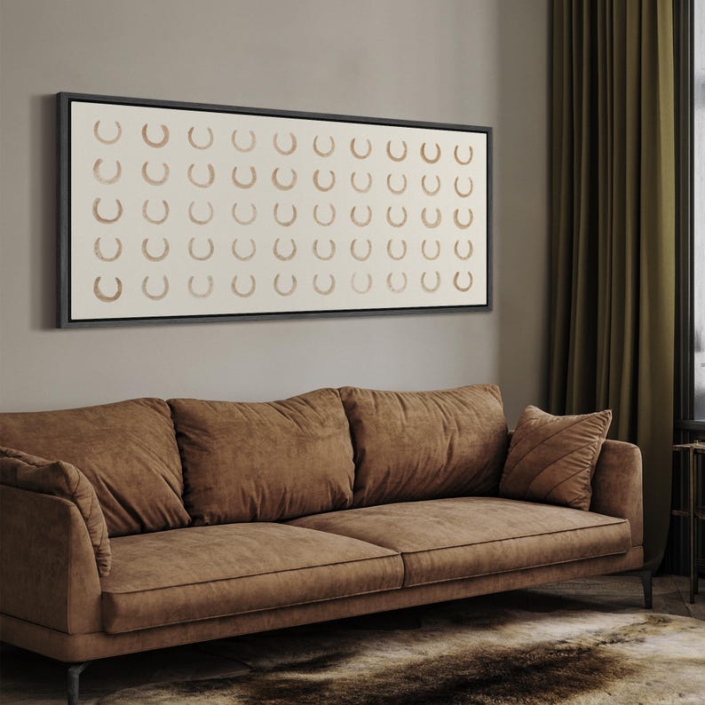May include: A large rectangular framed artwork featuring a repeating pattern of brown horseshoe shapes on a cream background. The artwork hangs above a brown sofa with matching throw pillows. The room has olive green curtains and a patterned rug.