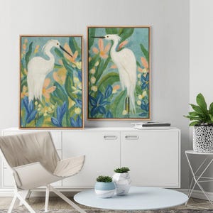 May include: Two framed watercolor paintings of white egrets in a lush, colorful garden setting. The artwork features blue, yellow, and peach flowers and foliage. The frames are a light wood color, and the paintings are displayed on a white cabinet.