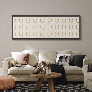 May include: A framed canvas art piece featuring a repeating pattern of gold horseshoes on a cream background. The artwork hangs above a beige sofa with decorative pillows and a wooden coffee table. The room has a neutral color palette.