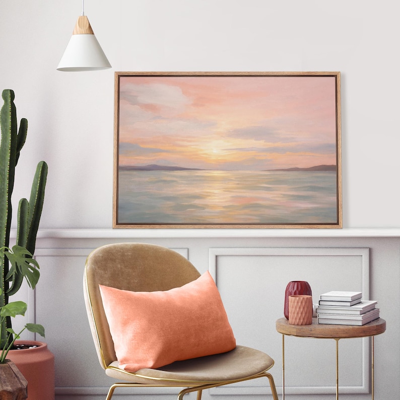 May include: A framed landscape painting depicts a serene sunset over water, with soft pink and yellow hues in the sky. A peach-colored pillow rests on a gold-legged chair. A small side table holds books and a decorative vase.