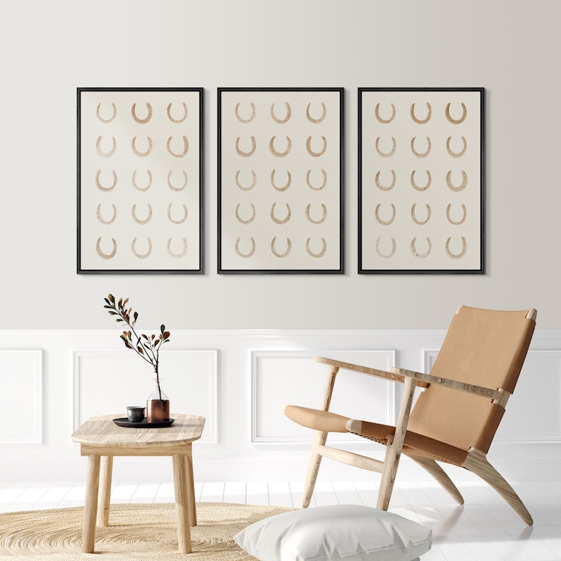 May include: Set of three framed art prints. Each print features a repeating pattern of tan horseshoes on a cream background. The frames are black. The artwork is displayed in a modern living room setting.