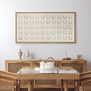 May include: A framed canvas art piece featuring a repeating pattern of tan horseshoes on a cream background. The artwork is displayed above a wooden cabinet with decorative items and a dining table with a tea set.