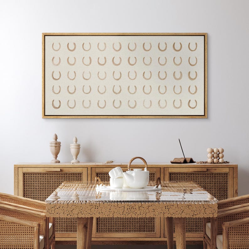 May include: A framed canvas art piece featuring a repeating pattern of tan horseshoes on a cream background. The artwork is displayed above a wooden cabinet with decorative items and a dining table with a tea set.