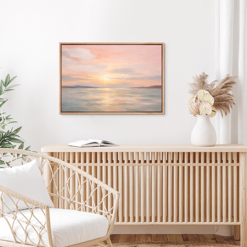May include: A framed landscape painting depicts a serene seascape at sunset. The sky is a blend of pink, orange, and yellow hues, reflecting on the water. The painting is in a natural wood frame.