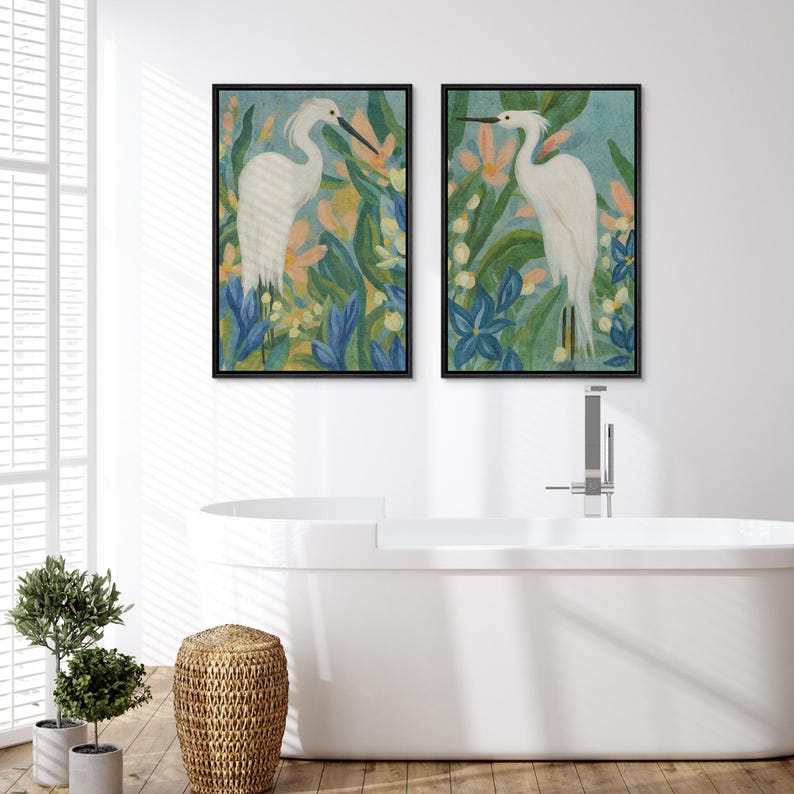 May include: Two framed art prints featuring white herons amidst colorful flowers and foliage. The artwork has a watercolor style with a light blue background, and the frames are black. The prints are displayed in a bathroom setting.