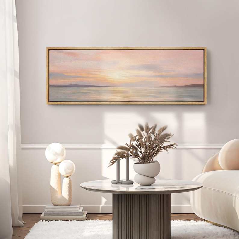May include: A framed landscape painting depicting a sunset over the ocean. The artwork features soft pastel colors of pink, orange, and blue. The frame is a light wood color, and the painting is displayed on a neutral-colored wall.