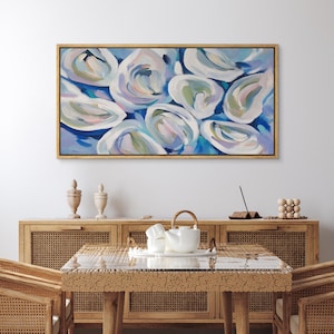 May include: Abstract painting of oyster shells in shades of white, blue, and green, set against a blue background. The artwork is framed in a gold-colored wooden frame. The painting hangs above a wooden cabinet and table.