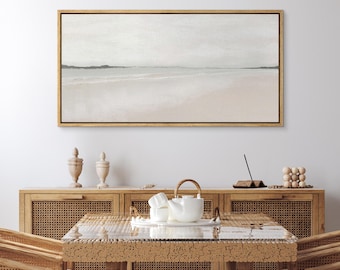 Puede incluir: Una pintura abstracta enmarcada que representa una escena de playa en tonos apagados de gris, blanco y beige. La pintura está colgada en una pared blanca sobre una mesa de madera con una tetera blanca y dos urnas decorativas.