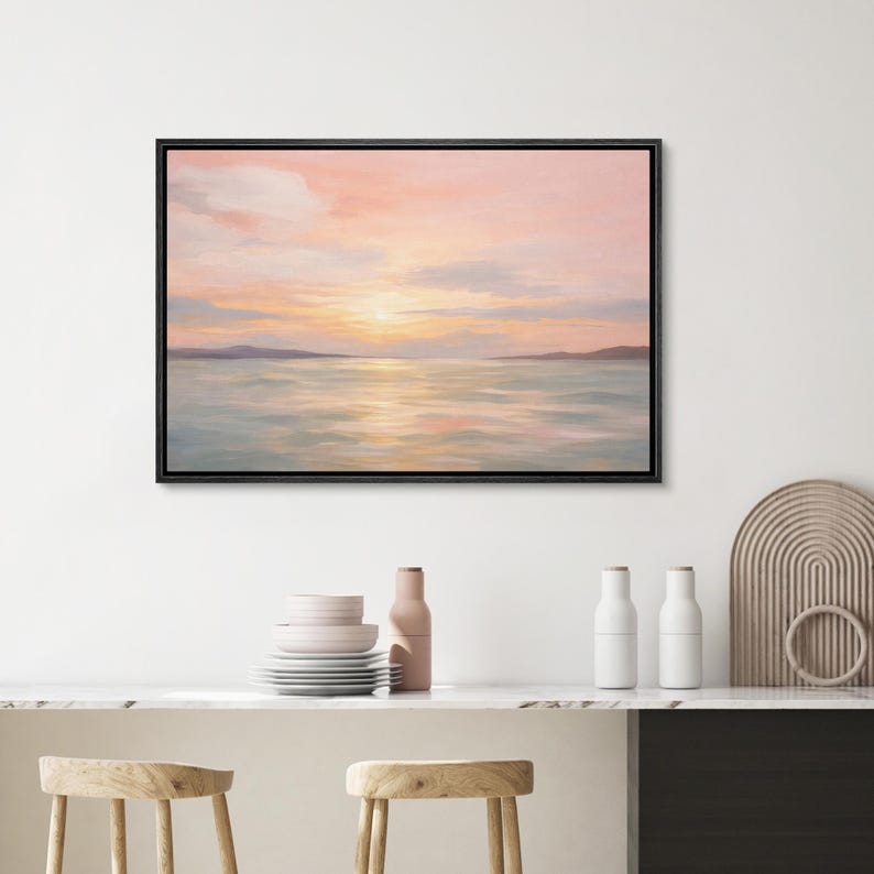 May include: A framed landscape painting depicts a serene sunset over water. The sky transitions from soft pink to yellow, reflecting on the water's surface. Distant landmasses are visible. The artwork is in a black frame.