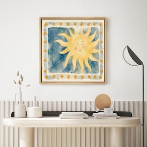 May include: A framed watercolor painting of a smiling sun with yellow rays on a blue background. The sun is centered within a square, surrounded by a border of yellow stars. The frame is a light brown color.