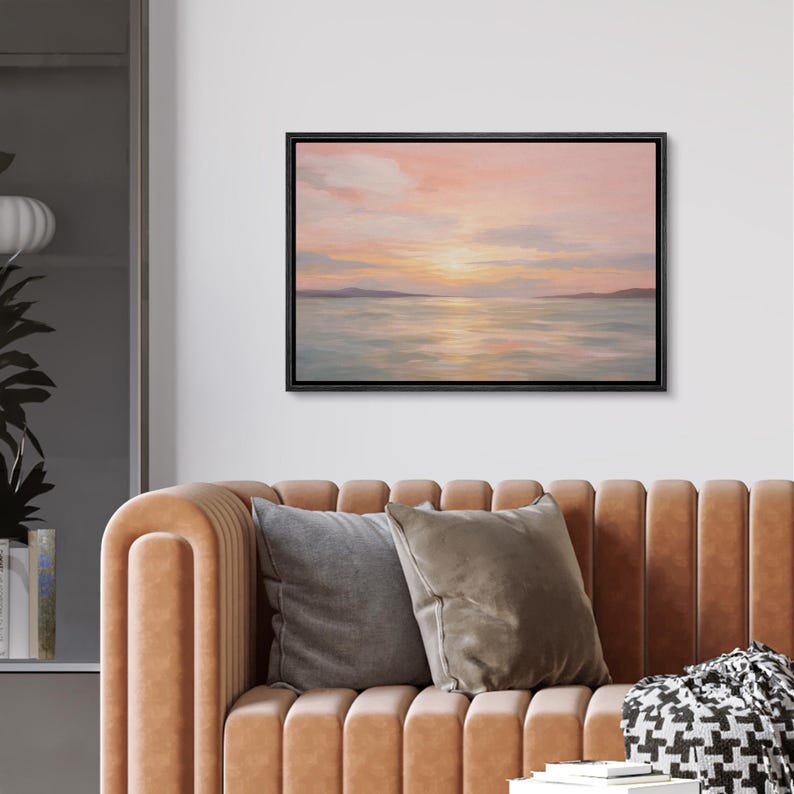 May include: A framed canvas art print depicting a serene seascape at sunset. The artwork features a soft color palette of pink, orange, and blue hues, with the sun reflecting on the water. The black frame complements the artwork.