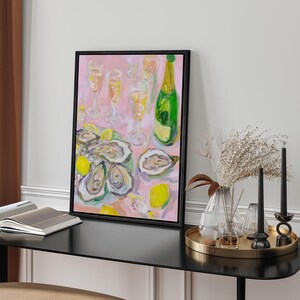 May include: A framed painting depicts a still life of oysters, champagne, and lemons on a pink background. A green champagne bottle and several glasses are visible. The painting is in a black frame and sits on a black table.