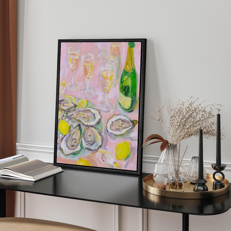 May include: A framed painting depicts a still life of oysters, champagne, and lemons on a pink background. A green champagne bottle and several glasses are visible. The painting is in a black frame and sits on a black table.