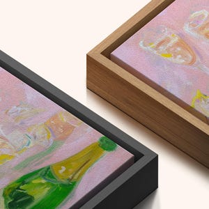May include: Two framed canvas art pieces. One features a green champagne bottle with a yellow label, the other shows champagne flutes. Both have a pink background. One frame is black, the other is natural wood.