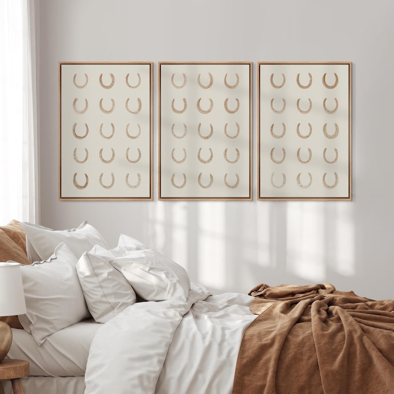 May include: Three framed wall art pieces. Each features a grid of tan horseshoes on a cream background. The frames are a light wood color. The artwork hangs above a bed with white linens and a brown blanket.