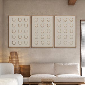 May include: Three framed wall art pieces. Each features a beige background with a repeating pattern of gold-colored horseshoes. The frames are a warm brown color. The artwork is displayed above a beige sofa and wooden table.