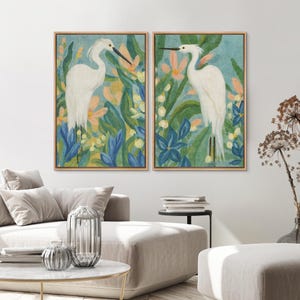 May include: Two framed art prints featuring white birds amidst colorful foliage. The artwork showcases a serene scene with blue, green, and peach tones, creating a calming aesthetic. The prints are displayed in wooden frames, adding a touch of natural warmth to the artwork.