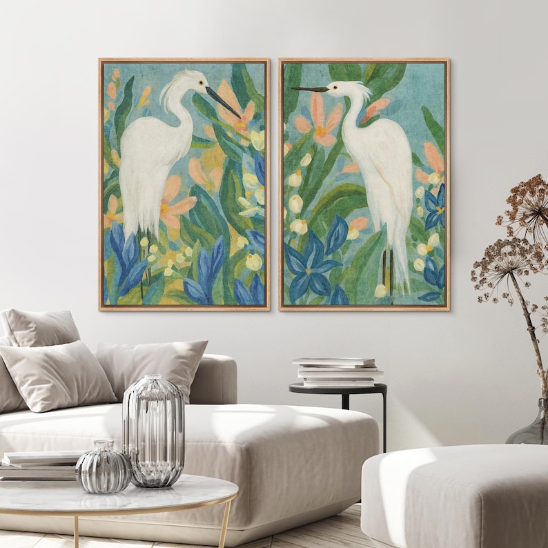 May include: Two framed art prints featuring white birds amidst colorful foliage. The artwork showcases a serene scene with blue, green, and peach tones, creating a calming aesthetic. The prints are displayed in wooden frames, adding a touch of natural warmth to the artwork.