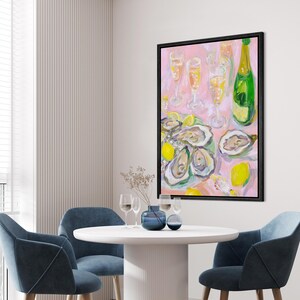 May include: A framed painting depicts a still life of oysters, lemons, champagne, and glasses on a pink background. The painting has a black frame and is displayed in a dining room with blue chairs and a white table.