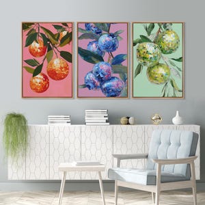 May include: Three framed art prints featuring colorful disco ball fruit. One shows orange disco balls on a pink background, another has blue disco balls on purple, and the last has green disco balls on a green background. The art is displayed above a white cabinet.