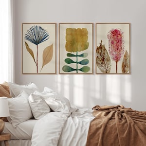 May include: A set of three framed wall art prints featuring botanical illustrations. The left print shows a blue fan-shaped flower with tan leaves. The center print displays a yellow block-like flower with green stems. The right print depicts a pink spiky flower.