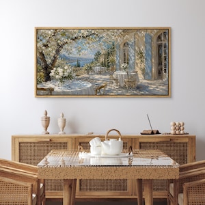 May include: A rectangular framed wall art print depicting a sunlit Mediterranean terrace with white flowering trees, tables set with white cloths, and blue shutters on a building, hanging above a wooden table with a white teapot and cups.