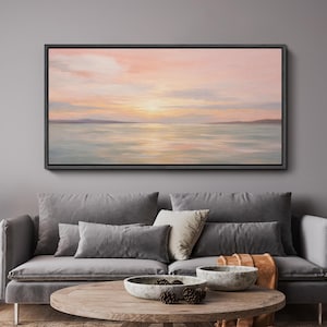 May include: A framed landscape painting depicts a serene seascape at sunset. The artwork features a soft color palette of pink, orange, and blue hues, with the sun's reflection on the water. The painting is displayed above a gray sofa.