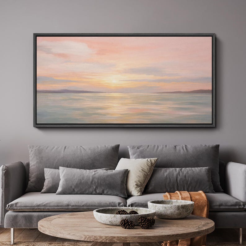 May include: A framed landscape painting depicts a serene seascape at sunset. The artwork features a soft color palette of pink, orange, and blue hues, with the sun's reflection on the water. The painting is displayed above a gray sofa.
