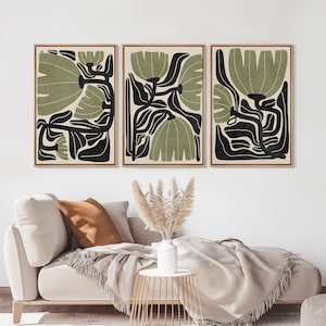 May include: A set of three rectangular wall art prints in light wood frames, each featuring a stylized botanical illustration with sage green and black organic shapes on a cream background, displayed in a horizontal row on a plain wall.