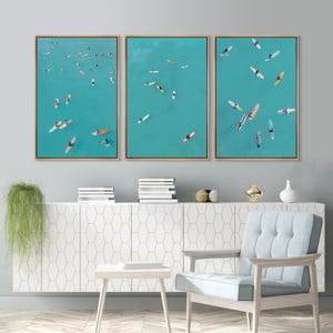 May include: A triptych of framed wall art featuring aerial views of people on colorful surfboards in turquoise ocean water. Each of the three vertical panels displays multiple individuals paddling on boards across the clear blue sea surface.