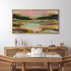 May include: Abstract landscape painting in a gold frame. The artwork features a serene scene with mountains, a lake, and colorful brushstrokes in shades of pink, green, and gold. The painting is displayed above a wooden cabinet.