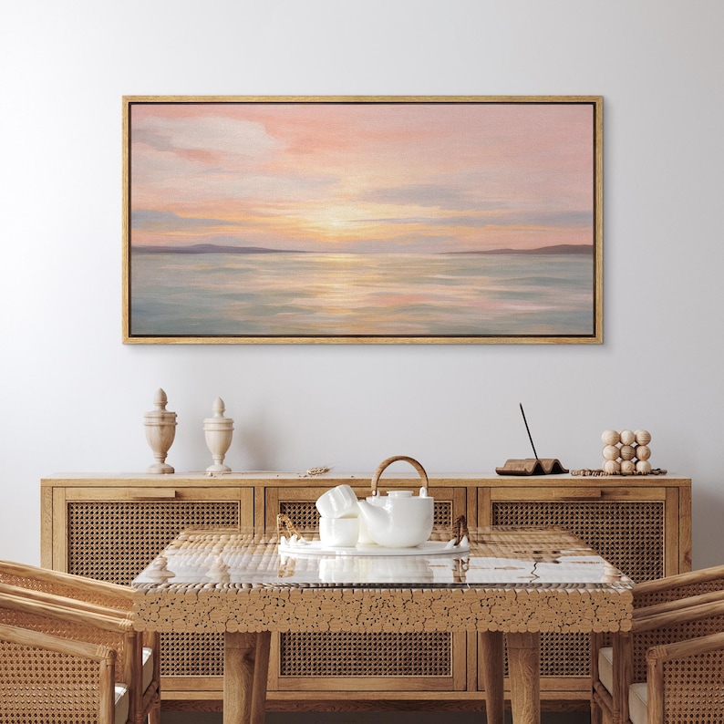 May include: A framed landscape painting depicts a serene sunset over a calm sea. The artwork features soft pastel colors of pink, orange, and blue, with a golden frame. The painting hangs above a wooden cabinet and table.