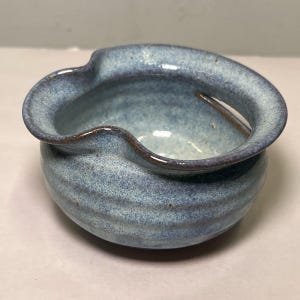 May include: A small, handmade ceramic bowl with a unique, wavy rim. The bowl is a speckled blue with darker blue accents and a brown rim. The exterior features horizontal ridges, adding texture to the piece. This decorative bowl is ideal for serving dips or small snacks.