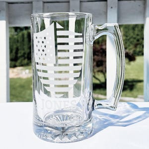 May include: A clear glass beer stein with a white shield design featuring an American flag pattern. The stein has a handle and the name "JONES" is etched on the front.
