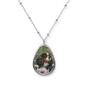 May include: Silver-toned pendant necklace featuring an oval charm with a Cavalier King Charles Spaniel portrait. The dog has a tricolor coat and is set against a backdrop of pink flowers and greenery. The chain is delicate and beaded.