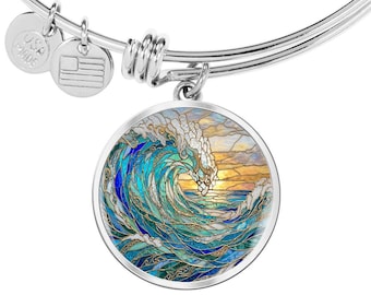 May include: A silver-tone expandable bangle bracelet featuring a round pendant with a stained glass style ocean wave design. The artwork displays swirling blue and teal water with white foam against a golden sunset sky. Two small metal charms are attached.