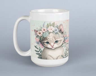 May include: White ceramic mug featuring a colourful illustration of a kitten wearing a flower crown. The kitten has blue eyes and is sitting on a green background with flowers.