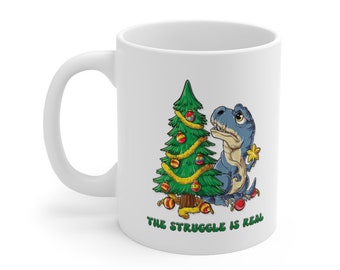 May include: White ceramic coffee mug with a blue dinosaur struggling to put a star on top of a Christmas tree. The text "THE STRUGGLE IS REAL" is printed below the image.