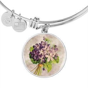 May include: Silver-toned bangle bracelet with a round charm featuring a vintage illustration of purple violets. The charm is accented with green leaves and tied with a gold-colored string. Includes two small USA-made charms.
