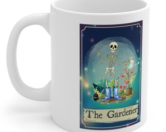 May include: White ceramic mug with a colorful illustration of a skeleton watering plants. The illustration is framed with a gold border and the text "The Gardener" is printed below the image.