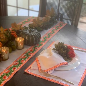 Può includere: Un runner da tavolo bianco con un design floreale verde e arancione. Il runner è su un tavolo in legno scuro con una mise en place bianca e un tovagliolo bianco. Ci sono anche due portacandele dorati e una zucca verde sul tavolo.