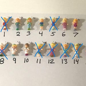 Può includere: Una collezione di quindici bambole in miniatura di plastica disposte in due file su una superficie bianca. Ogni bambola è numerata da 1 a 15, con alcune figure contrassegnate da una 'X' blu. Le bambole presentano vari colori di capelli e abiti.