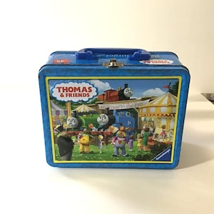 May include: A blue metal lunchbox featuring Thomas the Tank Engine and friends. The front of the lunchbox has a colorful illustration of the characters at a fair, with the words "Thomas & Friends" prominently displayed.