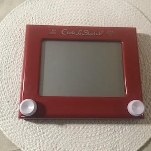 以下が含まれることがあります： 白いノブと上部に「1960 2020 Etch A Sketch 60」の文字が書かれた赤いプラスチック製のエッチ・ア・スケッチのおもちゃです。