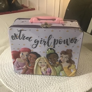 May include: A purple metal lunchbox with a pink handle and a white background. The lunchbox has a picture of Disney princesses on the front with the text "extra girl power".