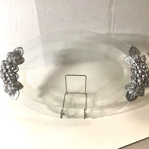 May include: An oval clear glass serving tray with a textured surface and two silver-toned metal handles shaped like clusters of grapes with leaves. The tray is supported by a thin, silver-colored wire stand that holds it in an upright position.