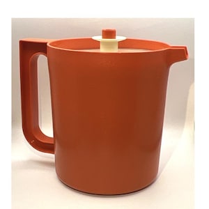 May include: An orange plastic pitcher with a handle and a spout. The pitcher has a lid with a white and orange knob. The pitcher is a solid color and appears to be vintage.