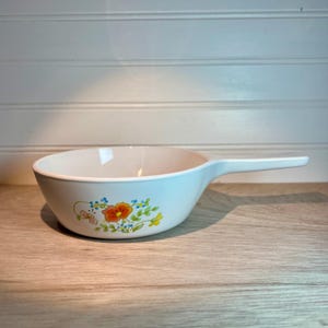 May include: White ceramic skillet with a long handle. The skillet features a floral design with orange, blue, and yellow flowers. The skillet is on a wooden surface.