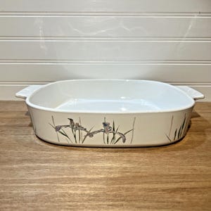 May include: Off-white rectangular baking dish with a floral iris design. The dish has a white interior and two handles. The iris design features purple and green accents. The dish is on a wooden surface.