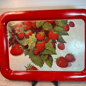 May include: A rectangular red metal serving tray with rounded corners. The tray features a painted image of ripe red strawberries with green leaves against a white background. The tray is supported by a black metal stand.